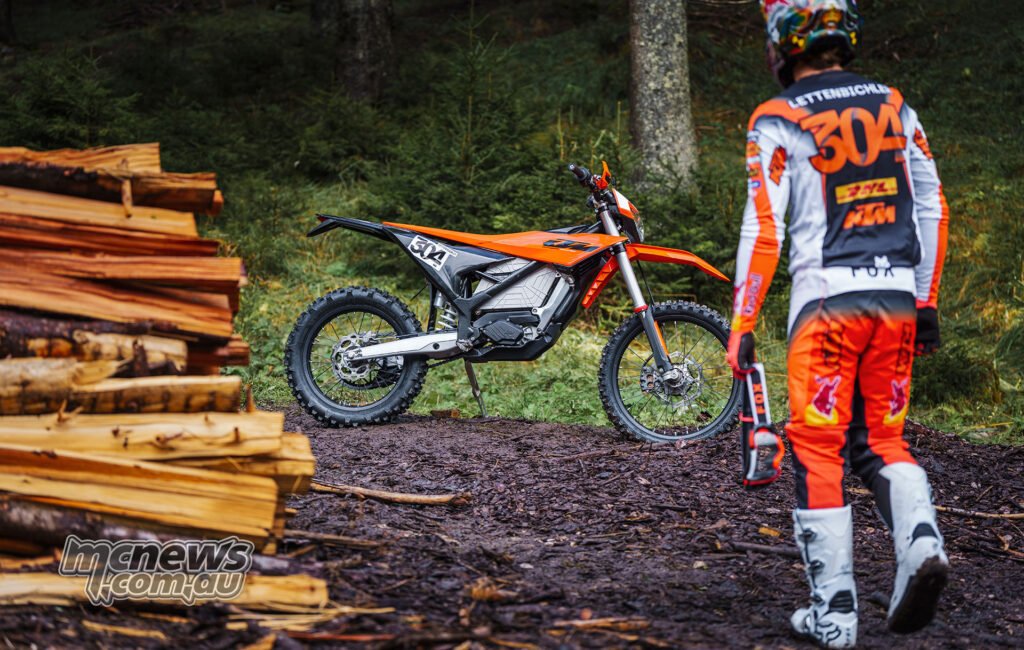 2027 KTM Freeride E parked on a forest trail with enduro star Mani Lettenbichler walking toward it.